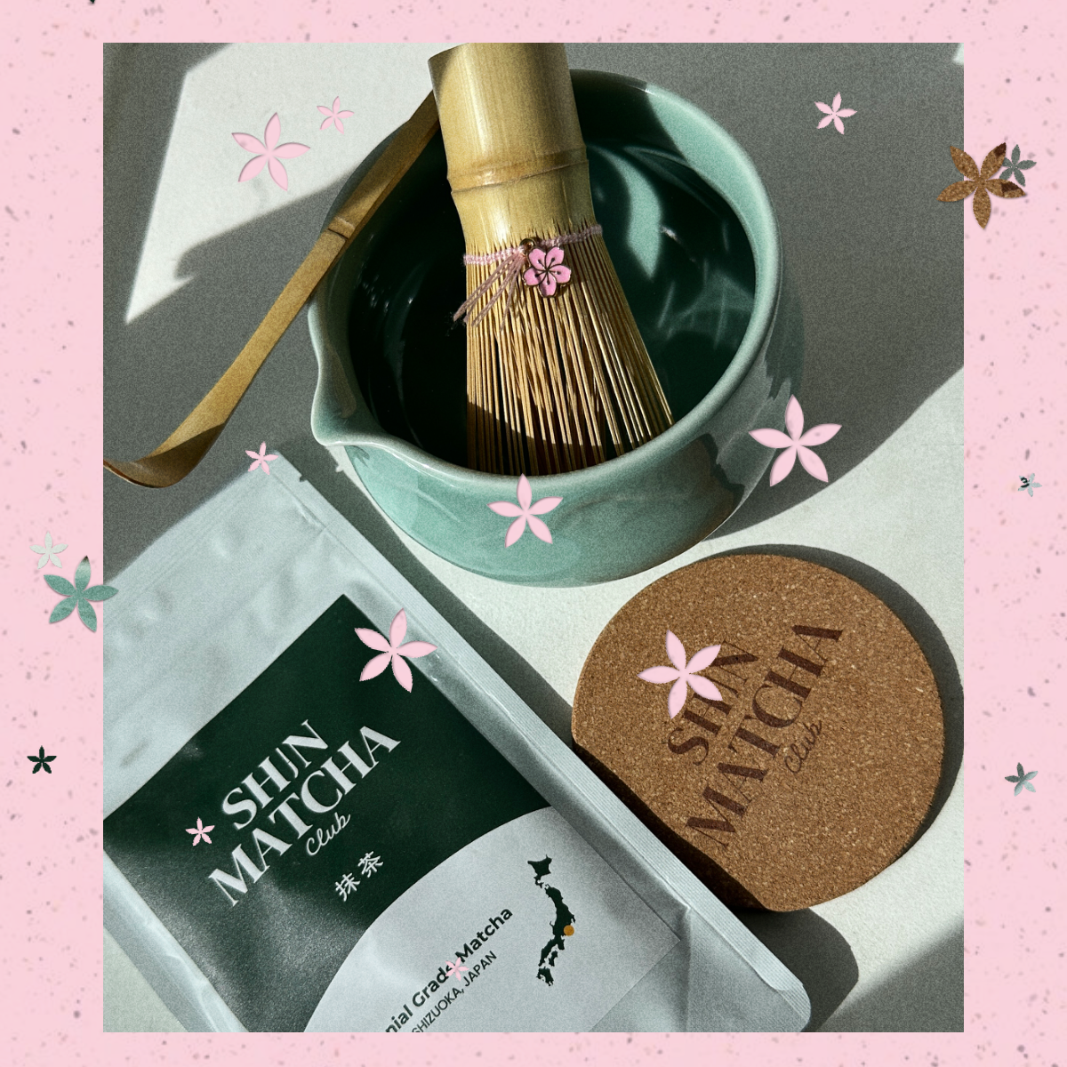 A styled flat-lay of a Shin Matcha tea product set on a pink floral background. The set includes a silver foil pouch of Shin Matcha Oolong tea, a round cork coaster with the brand name, a bamboo whisk in a light green ceramic bowl, and a bamboo scoop resting on the bowl.