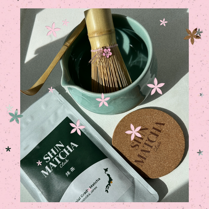 A styled flat-lay of a Shin Matcha tea product set on a pink floral background. The set includes a silver foil pouch of Shin Matcha Oolong tea, a round cork coaster with the brand name, a bamboo whisk in a light green ceramic bowl, and a bamboo scoop resting on the bowl.
